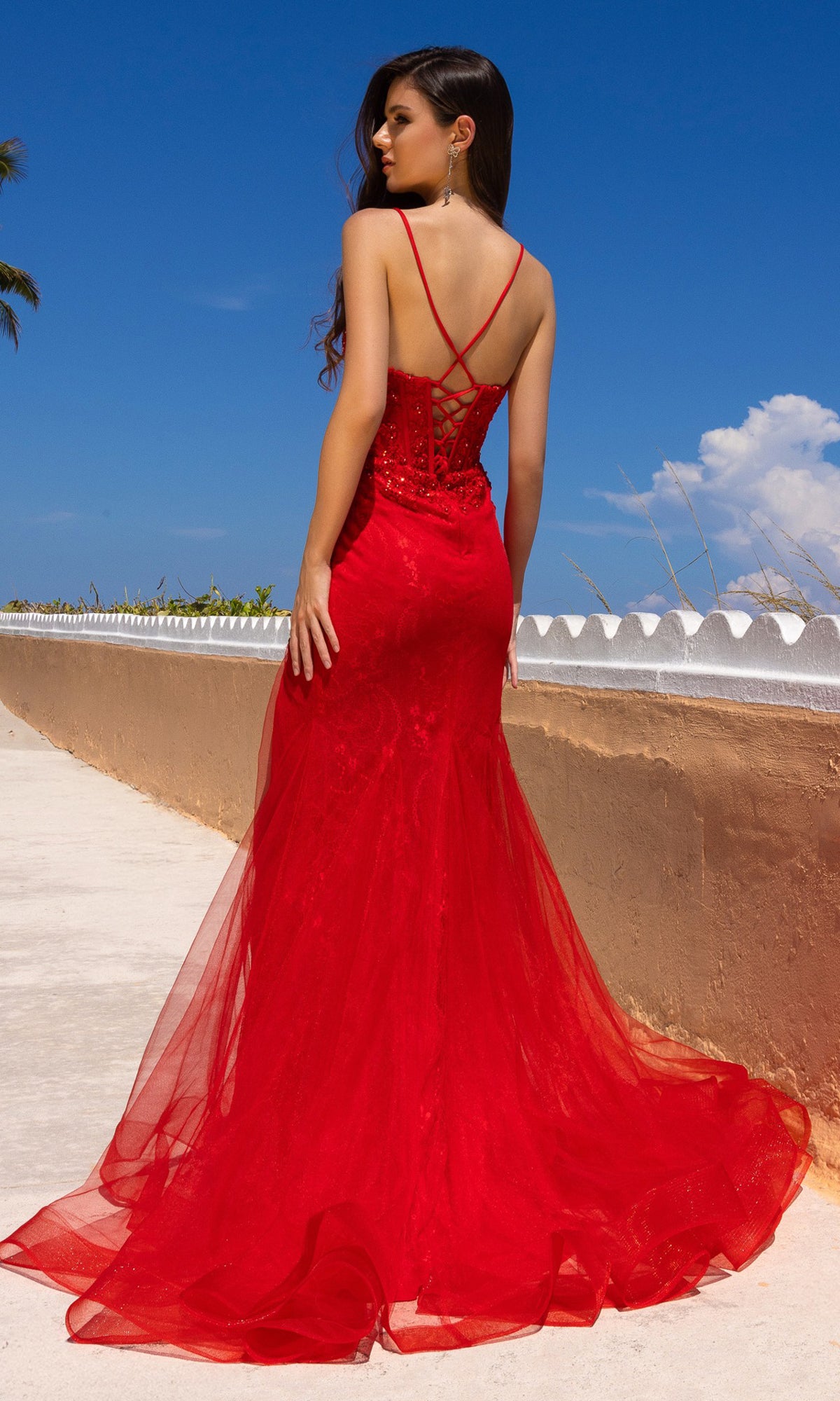 A woman stands outdoors on a sunny day, facing away from the camera, wearing the Amelia Couture 7064 Long Mermaid Prom Dress—a fitted red floral lace gown with thin straps and a lace-up back.